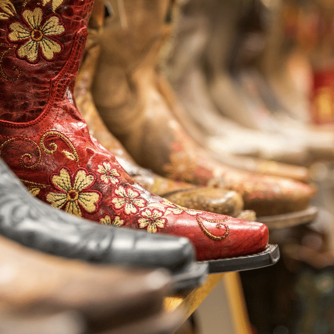 A close-up of a bright red cowboy boot with yellow floral embroidery, displayed among other brown and tan cowboy boots on a shelf in a store.