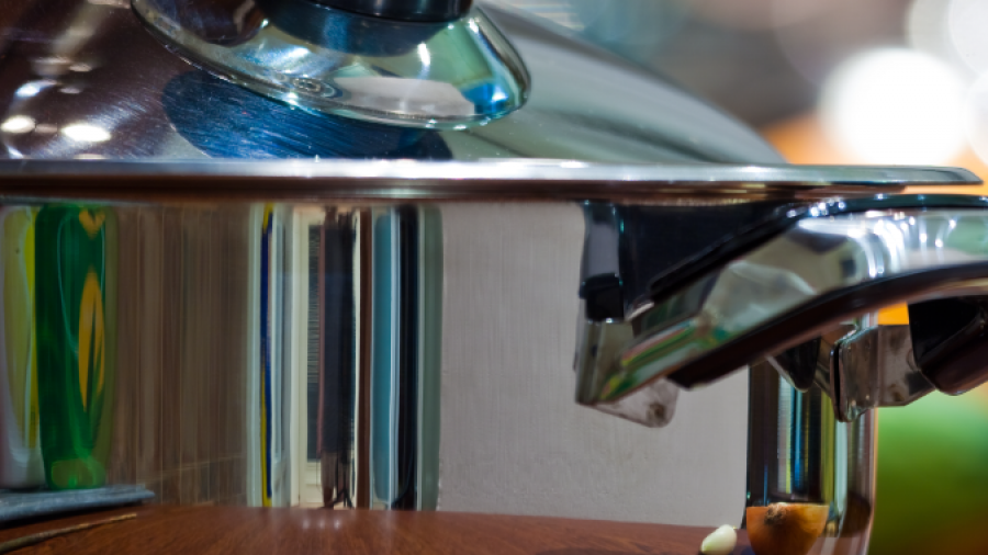 A close-up of a shiny stainless steel pot with a glass lid on a wooden surface. The pots handle and lid knob are visible, and reflections can be seen on the metal surface.