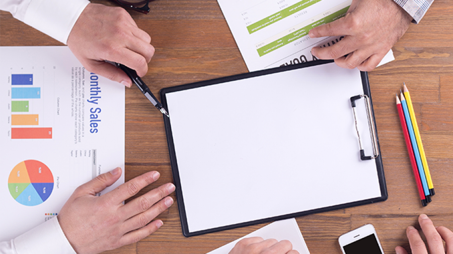 Four people sit around a wooden table with charts, graphs, a clipboard, pencils, glasses, a phone, and coffee, discussing business or sales data in a meeting.