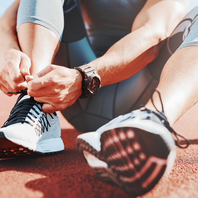 A person sitting on a running track ties the laces of a white and black athletic shoe. They wear sportswear and a watch, preparing for physical activity under bright sunlight.