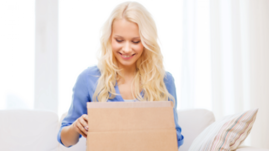 A smiling woman with long blonde hair sits on a couch and opens a cardboard box, looking inside with excitement. Light streams in from large windows behind her.