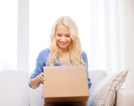 A smiling woman with long blonde hair sits on a couch and opens a cardboard box, looking inside with excitement. Light streams in from large windows behind her.