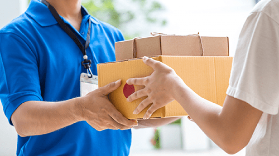 A delivery person in a blue shirt hands multiple packages, including a large yellow box, to another person. Both faces are out of frame, focusing on the exchange of parcels.