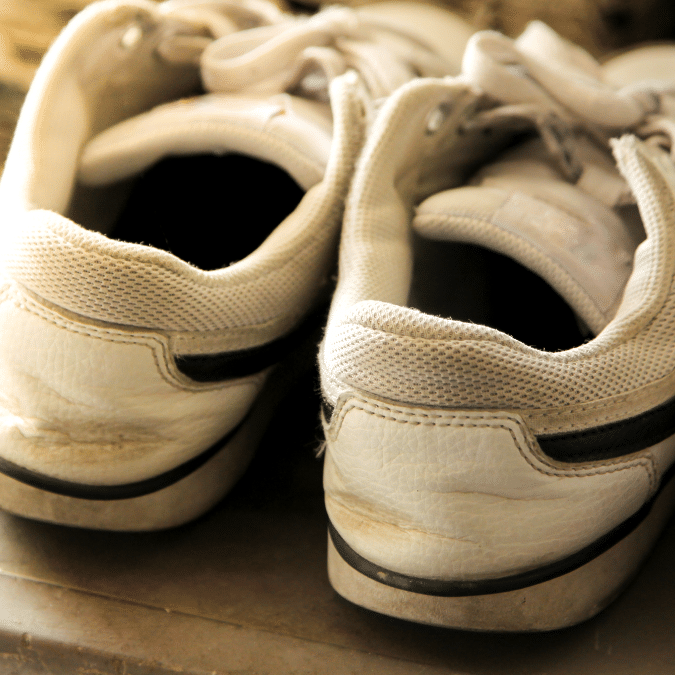 A close-up of the back of a pair of white sneakers with black accents, showing signs of wear and light dirt marks, placed side by side on a surface.