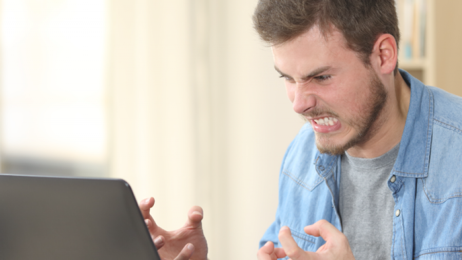 A man sitting at a desk in front of a laptop clenches his fists and grimaces in frustration. Papers, a cup, and a smartphone are on the desk, and bookshelves are in the background.