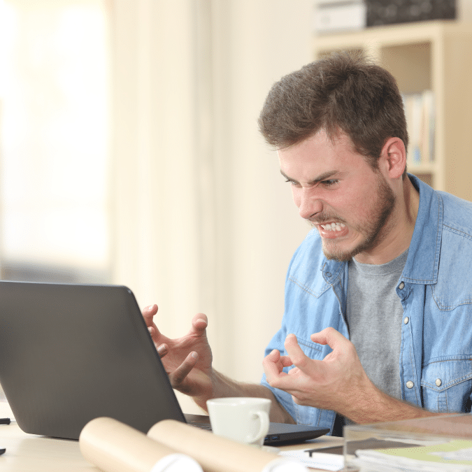 A man sitting at a desk in front of a laptop clenches his fists and grimaces in frustration. Papers, a cup, and a smartphone are on the desk, and bookshelves are in the background.