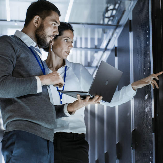 Two people in business attire, wearing lanyards, stand in a server room. One holds a laptop while the other points at a server rack, both focused and engaged in work-related discussion.