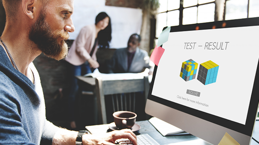 A man with a beard works on a desktop computer in a modern office. His screen displays a test result with images of colored cubes. Two colleagues are blurred in the background at a table.