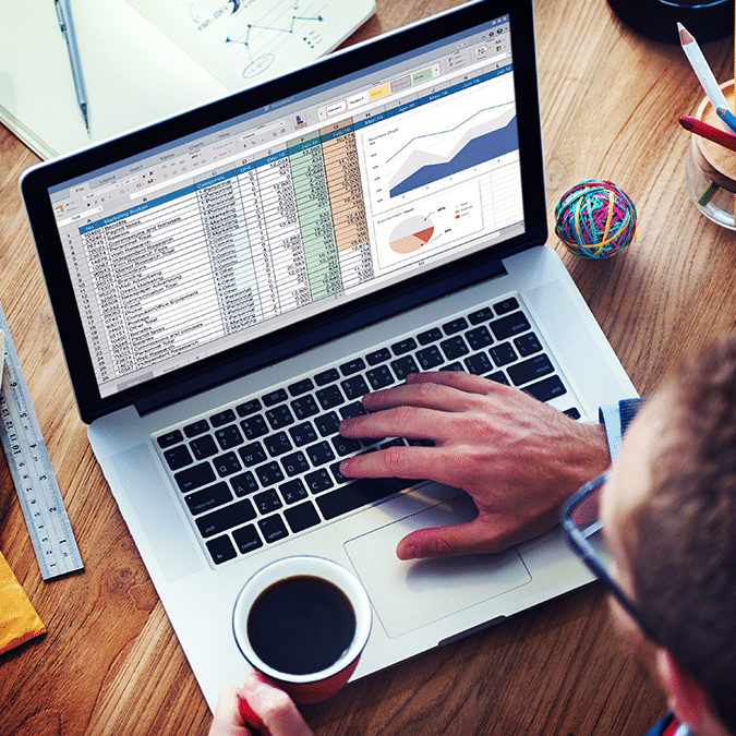 A person holding a cup of coffee works on a laptop displaying a spreadsheet with data tables, charts, and graphs. Office supplies, a notebook, and a colorful rubber band ball are on the wooden desk.