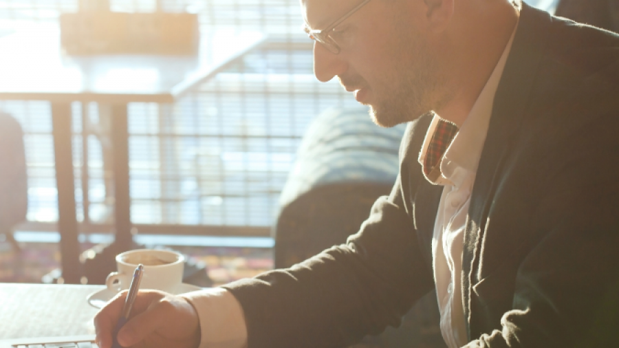 A man in a suit sits at a desk with a laptop, writing on paper with a pen. Sunlight streams through a window behind him, and a cup of coffee is on the table.