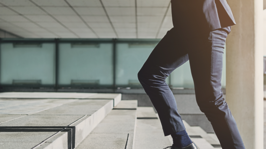 A person in a business suit walks up a set of outdoor concrete stairs, with only their lower body visible. The background features modern architecture and large windows.