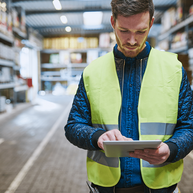 A man wearing a yellow safety vest uses a digital tablet in a warehouse aisle, focusing on the screen with shelves and industrial lighting in the background.