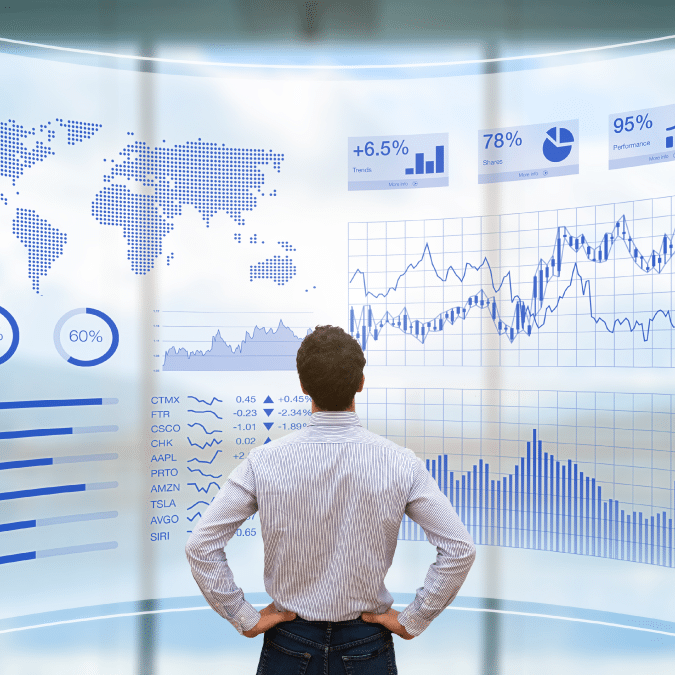 A man stands facing a large digital screen displaying global maps, charts, and financial graphs with data, percentages, and stock information, suggesting financial analysis or business data visualization.
