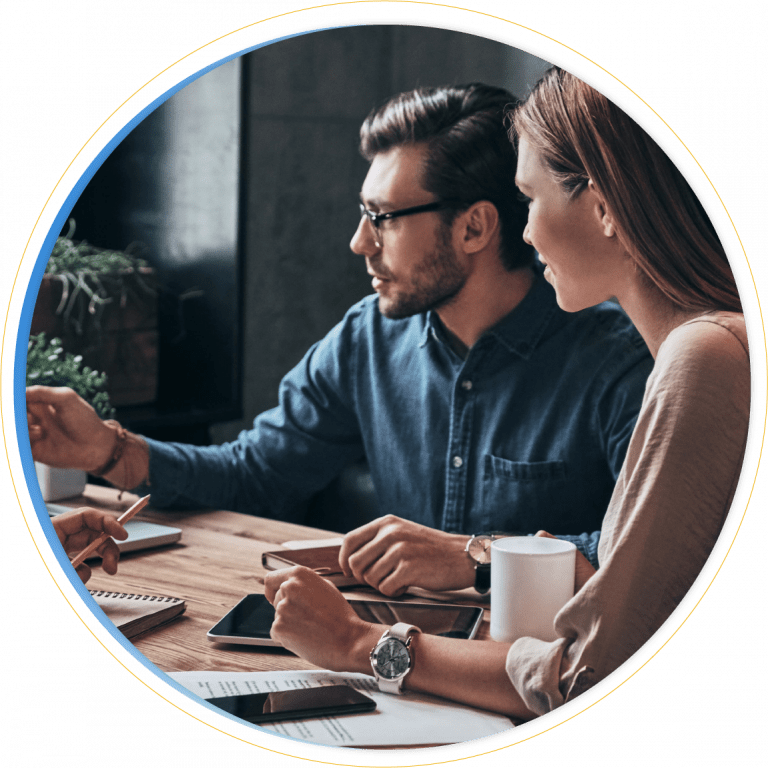 Two people sit at a wooden table working together, looking at a computer screen with Dynamics 365 Marketing open. One holds a pen and paper, while the other gestures toward the screen. A smartphone, notebook, and coffee mug are also on the table.