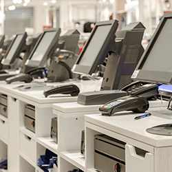 A row of empty retail checkout counters with touchscreen monitors, payment terminals, receipt printers, and pens, in a bright store environment.