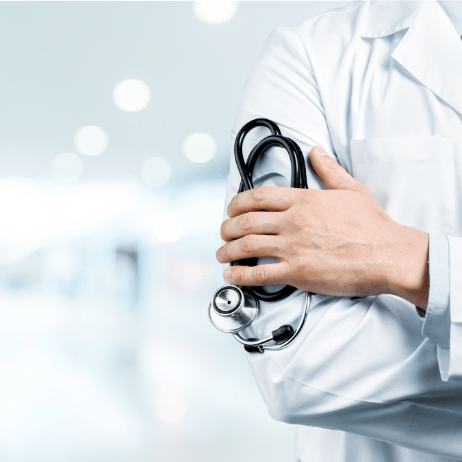 A doctor in a white coat stands with arms crossed, holding a stethoscope, in a bright, blurred medical or hospital setting.