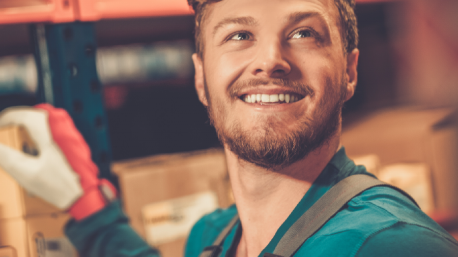 A smiling man wearing work gloves and an apron is organizing boxes on shelves in a warehouse or storage area, looking up with a cheerful expression.
