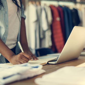 A person stands at a table, writing in a notebook with one hand while the other rests nearby. An open laptop, folded clothes, and blurred clothing racks suggest an environment focused on Retail Operations Consulting.