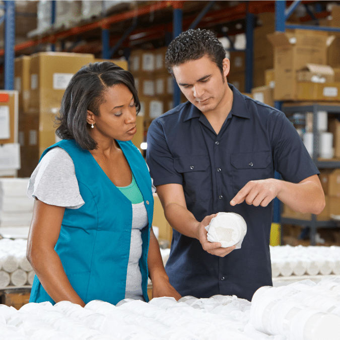 Two workers stand in a warehouse, examining a cylindrical product. The man points at details on the item while the woman listens attentively. Shelves filled with boxes and products are visible in the background.