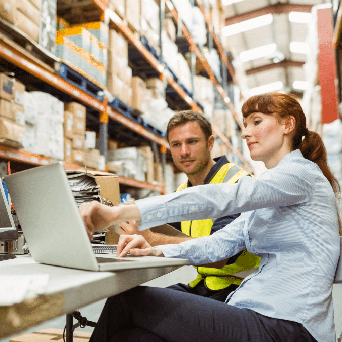 Two workers in a warehouse sit at a table with a laptop, discussing something on the screen. One wears a safety vest and the other is dressed in business attire. Shelves stocked with boxes are visible in the background.