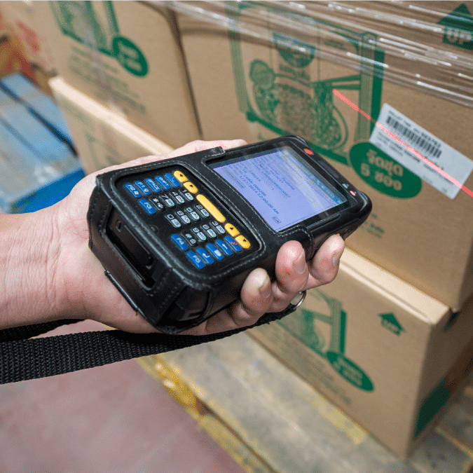 A person uses a handheld barcode scanner to scan a barcode on a cardboard box in a warehouse setting.