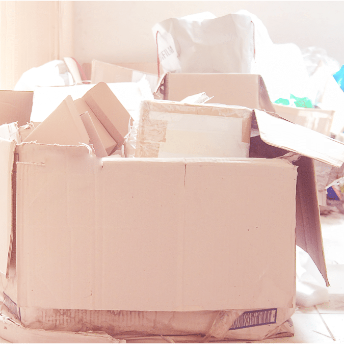 A large, open cardboard box filled with various smaller boxes and packing materials sits on a tiled floor in a brightly lit room. Other boxes and bags are visible in the background.