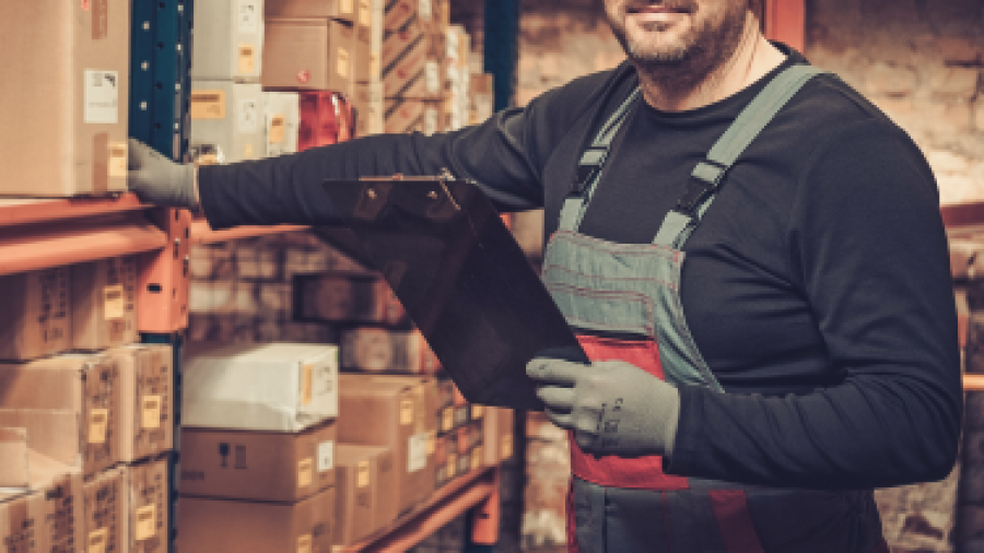 A warehouse worker in overalls and gloves stands by shelves of boxes, holding a clipboard and smiling at the camera in a well-lit storage area.