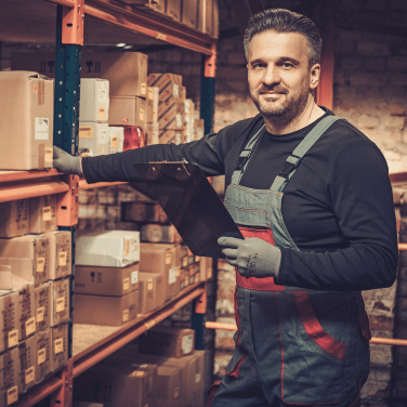 A warehouse worker in overalls and gloves stands by shelves of boxes, holding a clipboard and smiling at the camera in a well-lit storage area.