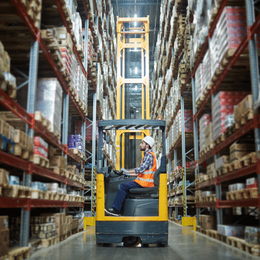 A worker wearing a hard hat and safety vest operates a forklift in a warehouse aisle lined with tall shelves stocked with various boxes and goods.