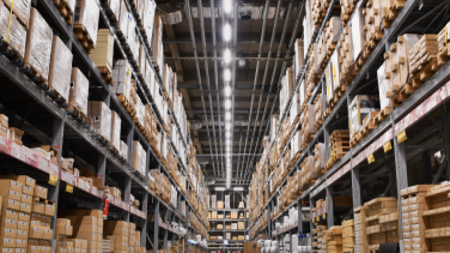 Wide-angle view of a large warehouse with tall shelves stacked with boxes, packages, and goods. The polished floor reflects the bright overhead lights, and the shelves extend into the distance.