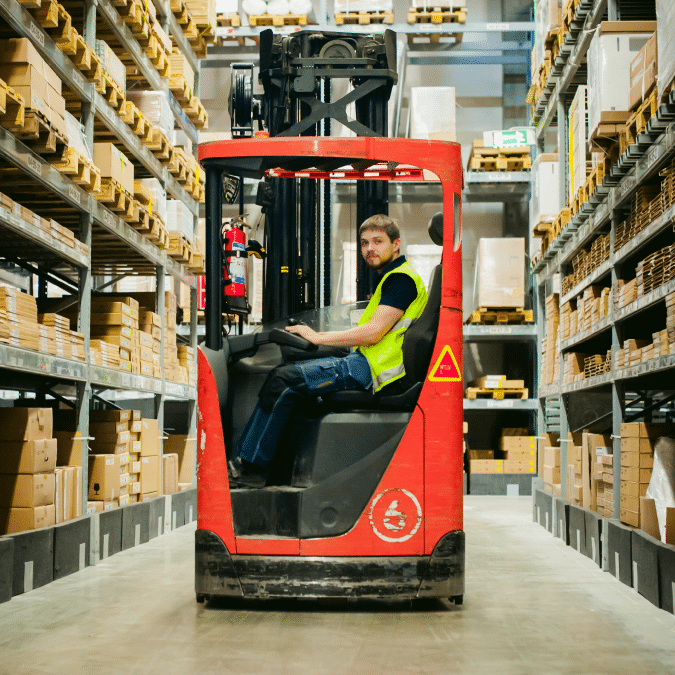 A man wearing a yellow safety vest drives a red forklift down an aisle in a warehouse, surrounded by shelves filled with cardboard boxes.