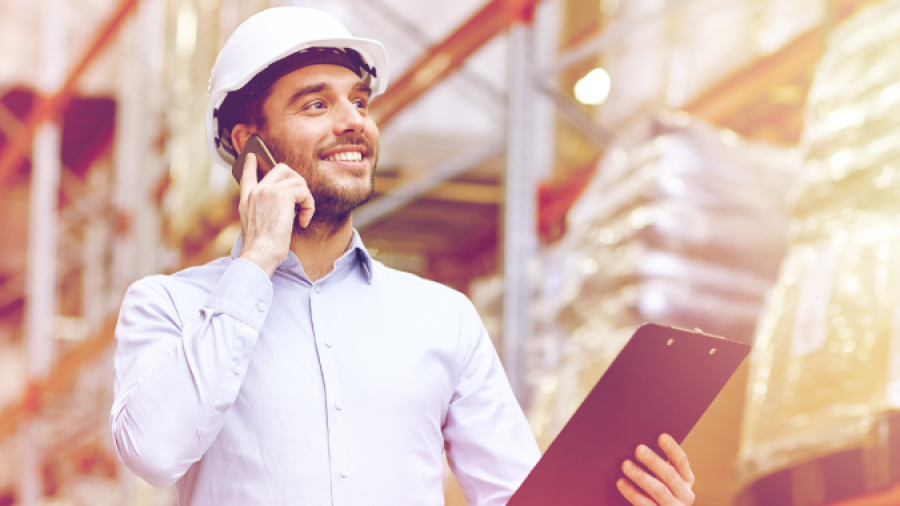 A man wearing a white safety helmet stands in a warehouse, smiling while talking on a phone and holding a clipboard. Tall shelves with boxes and packages are visible in the background.