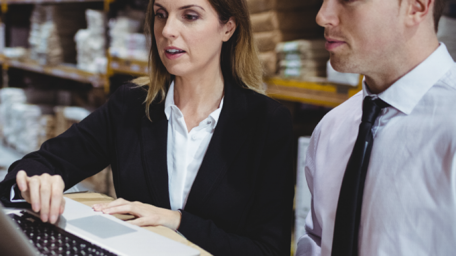 Two business professionals in formal attire look at a laptop on a desk in a warehouse, surrounded by shelves filled with boxes and packages.
