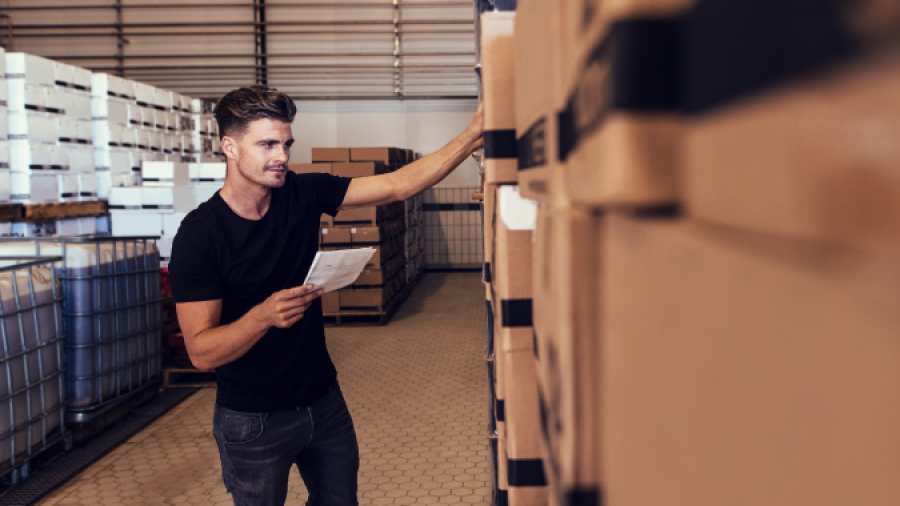 A man in a black t-shirt holds a checklist while examining stacked cardboard boxes in a warehouse, appearing to take inventory or check deliveries.