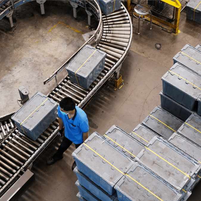 A person in a blue shirt and face mask walks beside a conveyor belt with large gray crates in an industrial warehouse setting. More crates are stacked neatly nearby.
