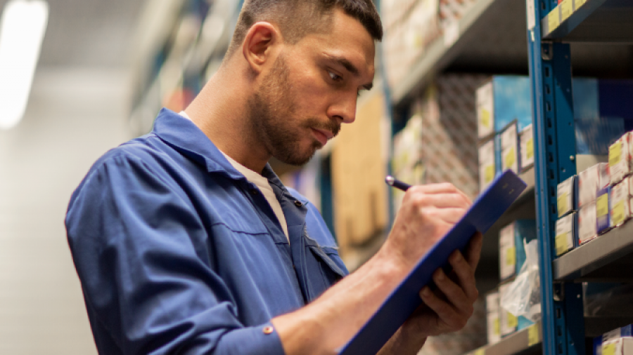 A man in a blue work uniform writes on a clipboard while standing in front of shelves filled with boxes in a storage or warehouse setting.