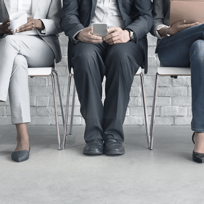 Three people in business attire sit on chairs against a white brick wall. They are holding phones or tablets, and only their bodies and legs are visible, not their faces.