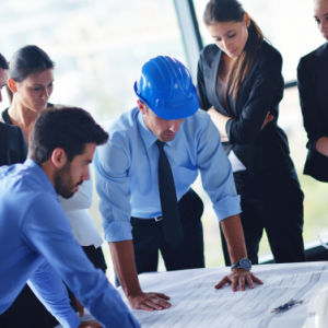 A group of business professionals, including one in a blue safety helmet, stand around a table reviewing architectural plans in a modern office—a scene reflecting project management consulting expertise.