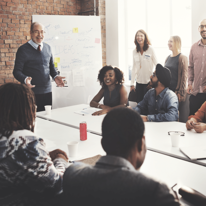 A diverse group of people are smiling and engaging in a meeting around a conference table, with one person standing and presenting ideas on a whiteboard in a modern office setting.