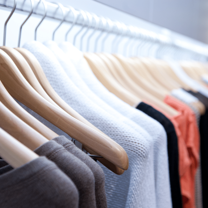 A row of assorted sweaters in neutral and muted colors hangs neatly on wooden hangers on a clothing rack.