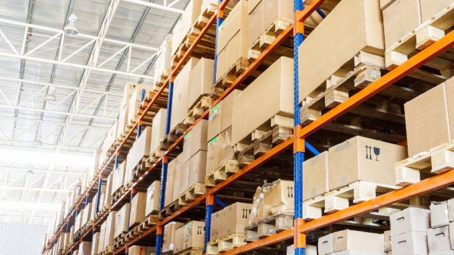 Large warehouse interior with tall shelves stacked with cardboard boxes and packages on wooden pallets. The space is brightly lit with natural and artificial light, and the shelves are organized and orderly.
