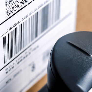 Close-up of a shipping label with a barcode on a cardboard box, next to a black scanner or device positioned for reading the barcode.