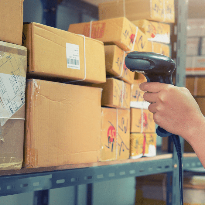 A person scans a barcode on a cardboard box with a handheld scanner in a warehouse, surrounded by stacked packages on metal shelves.