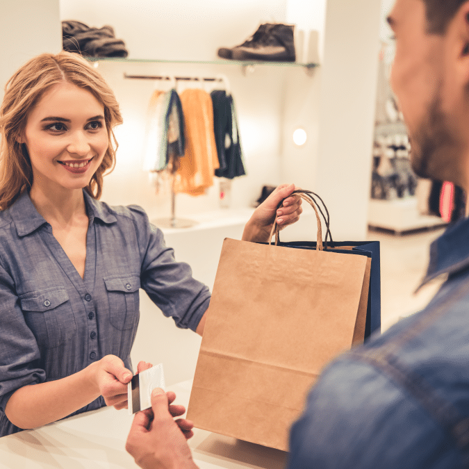 A smiling woman at a clothing store counter hands a paper shopping bag to a customer who is holding a credit card. Clothing racks and shelves are visible in the background.