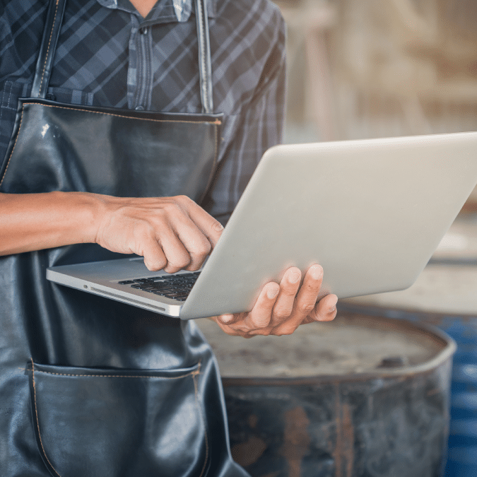A person wearing a dark apron and plaid shirt uses a laptop, standing in what appears to be a workshop or industrial setting. Only the torso and hands are visible.