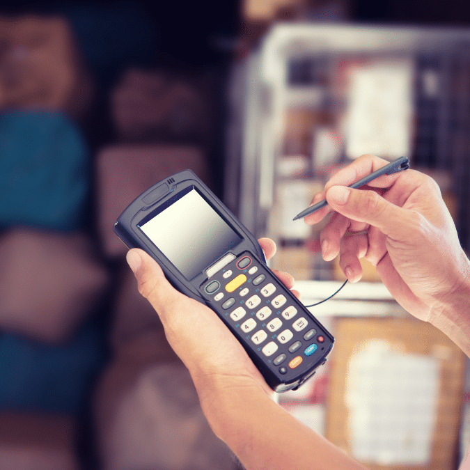 A person uses a stylus on a handheld barcode scanner or inventory device, with shelves and packages blurred in the background.