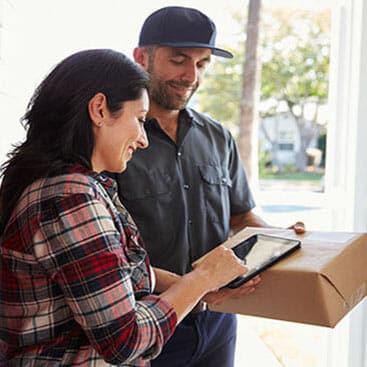 A woman signs for a package on a digital tablet held by a delivery person wearing a cap and uniform, standing at the doorway of a house.