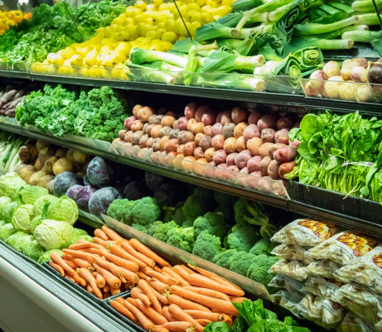 Fresh vegetables displayed on supermarket shelves, including lettuce, carrots, broccoli, cabbage, potatoes, leeks, yellow squash, and packaged goods, arranged neatly in rows for sale.