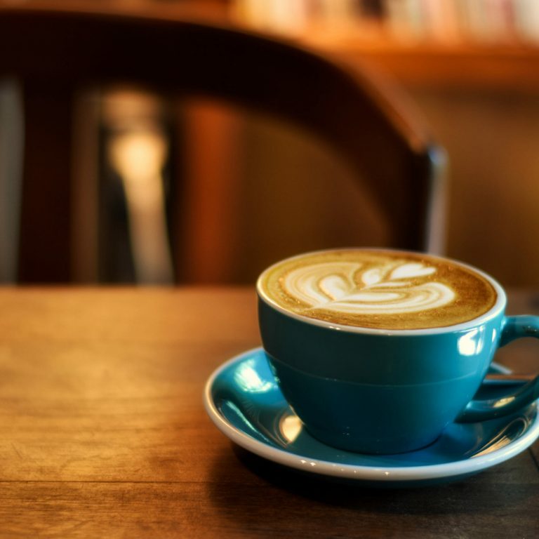 A blue cup of latte with intricate foam art sits on a matching saucer atop a wooden table, evoking the thoughtful ambiance often found in Business Intelligence Consulting spaces, with a blurred chair and bookshelf in the warm, softly lit background.