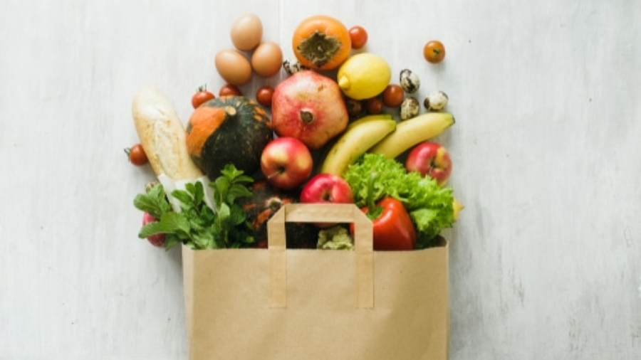 A paper grocery bag overflows with fresh produce, bread, leafy greens, apples, bananas, eggs, pomegranate, squash, tomatoes, strawberries, and other colorful fruits and vegetables against a light background.